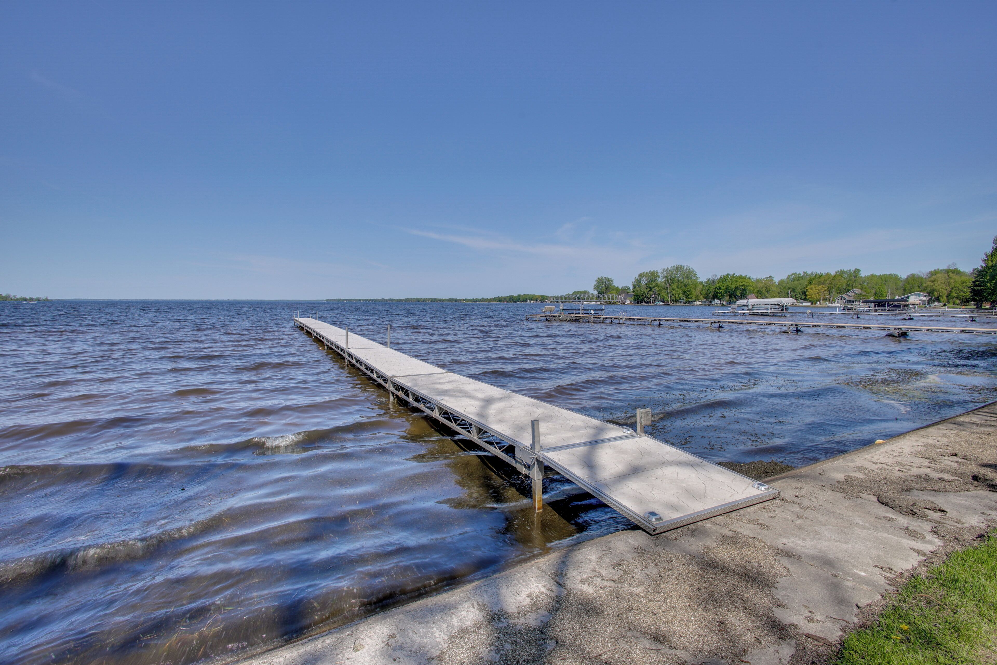 A concrete dock extending into calm water, a grassy area, and a distant shoreline with trees and buildings.
