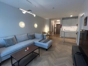 A modern living room with a sofa, a coffee table, and a dining area with chairs. There is a kitchen area with cabinets and a countertop. - Kalpaka Residence Apartment  (Rīga)