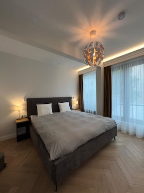 A modern hotel bedroom with a large bed, bedside tables, a ceiling light fixture, and large windows with curtains. - Kalpaka Residence Apartment  (Rīga)