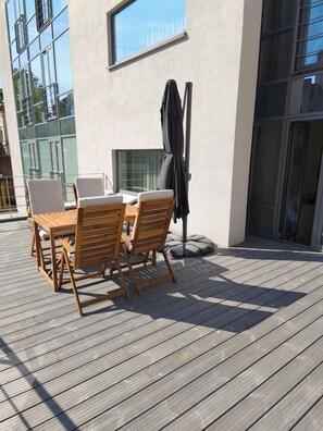 A wooden deck with a table and chairs, a covered grill, and a window overlooking a building. - Kalpaka Residence Apartment  (Rīga)