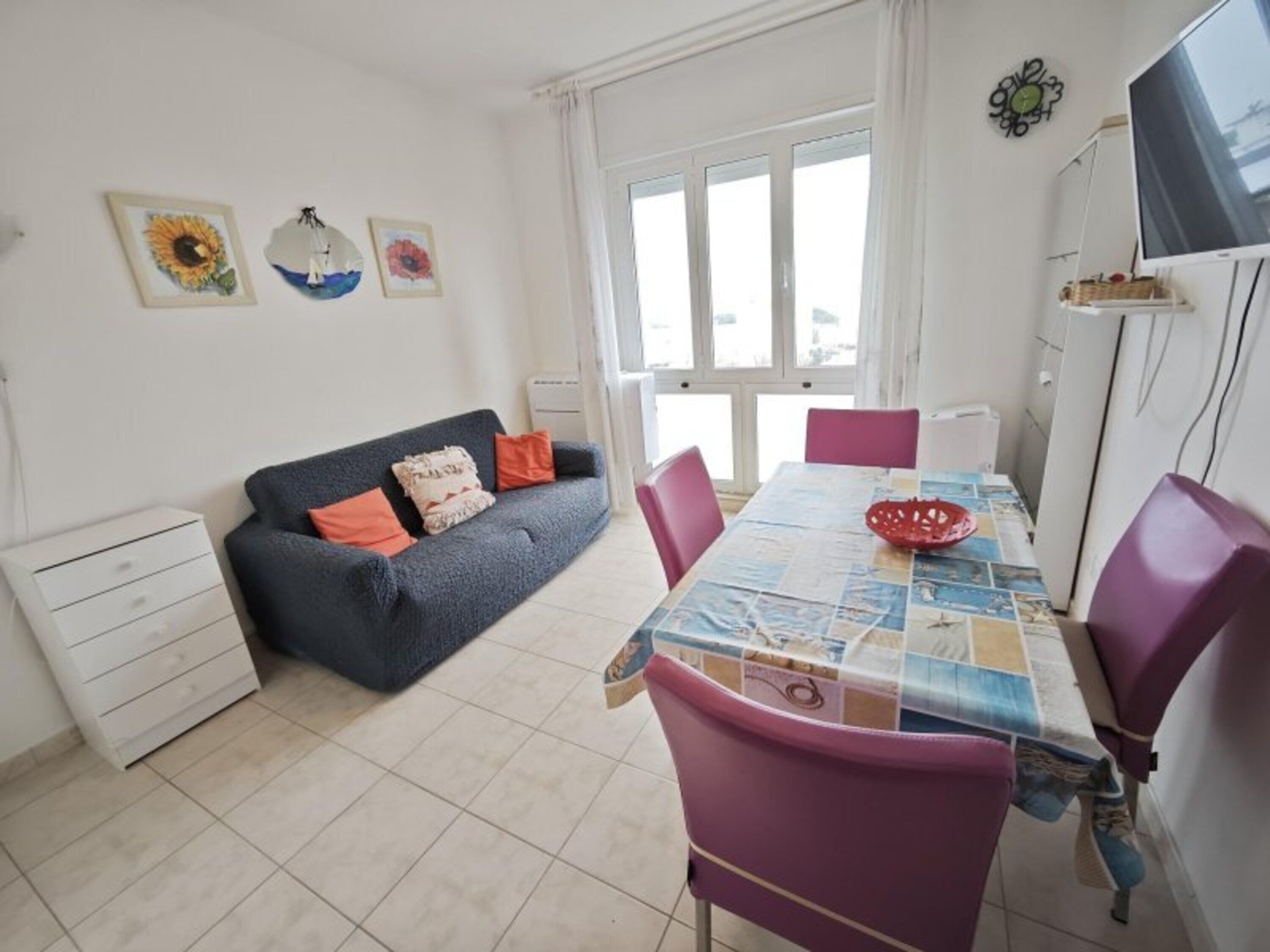 A dining area with a table set for two, a sofa, a TV, and a window with curtains.