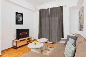 Ein Wohnzimmer mit Flachbildfernseher, rundem Couchtisch, einer Couch und einem großen Fenster mit Vorhängen.