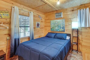 Ein Zimmer in einer Holzhütte mit einem Bett, einem Gemälde, einem Regal und einem Fenster mit Vorhängen.