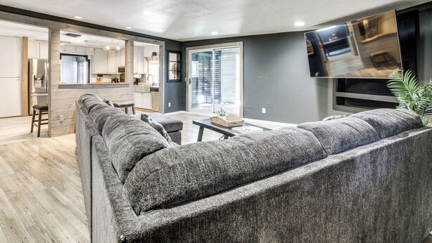 Ein modernes Wohnzimmer mit einem großen grauen Sofa, einem Wand-Fernseher und einer Küche im Hintergrund.
