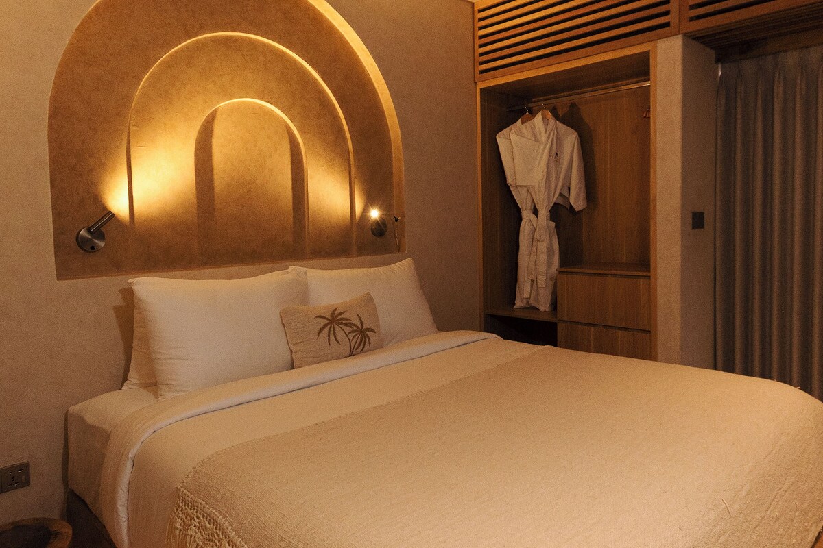 Una habitación de hotel con una cama, una bata colgada en un armario de madera y una lámpara de pared.
