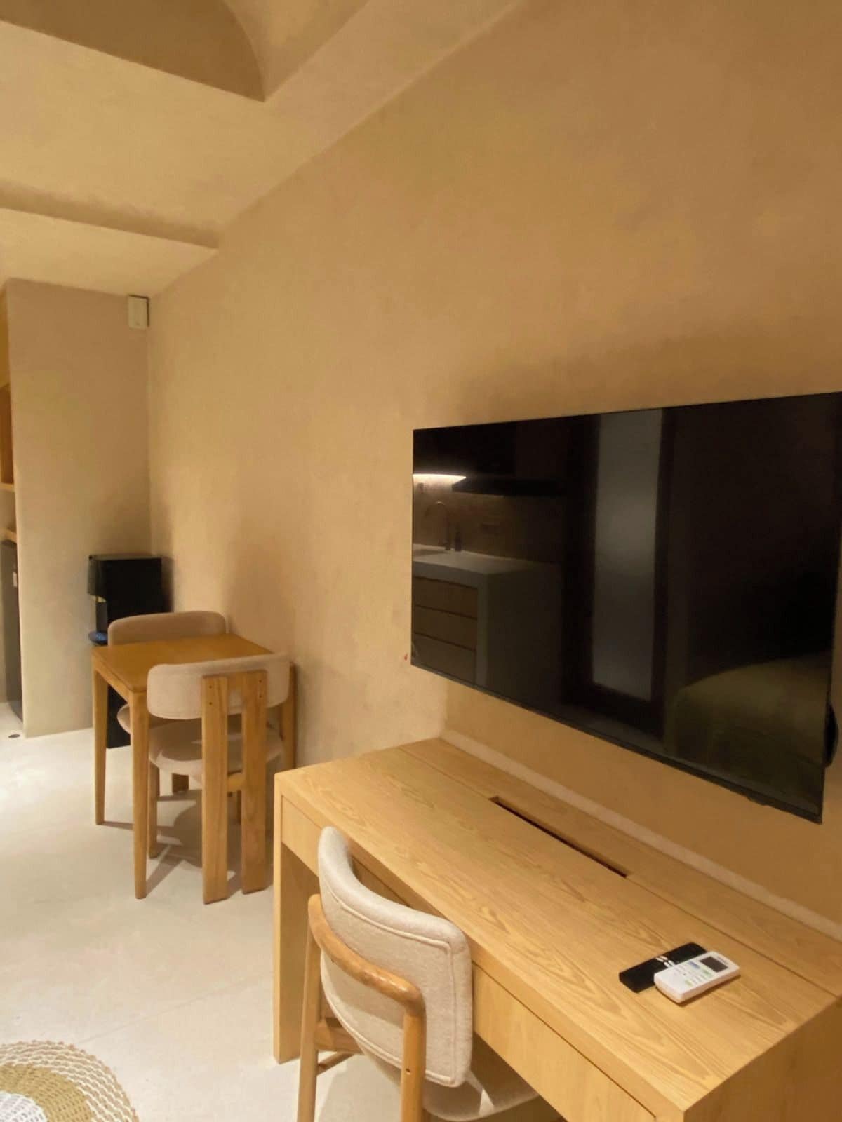 Una habitación moderna con un escritorio de madera, una silla, un televisor fijado a la pared y una mesita con un control remoto.