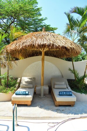 A poolside lounge area with two sunbeds, a thatched umbrella, and a palm tree in the background.