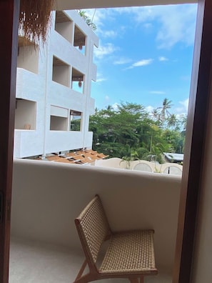 A balcony with a woven chair, a white building with multiple balconies, and tropical vegetation in the background.