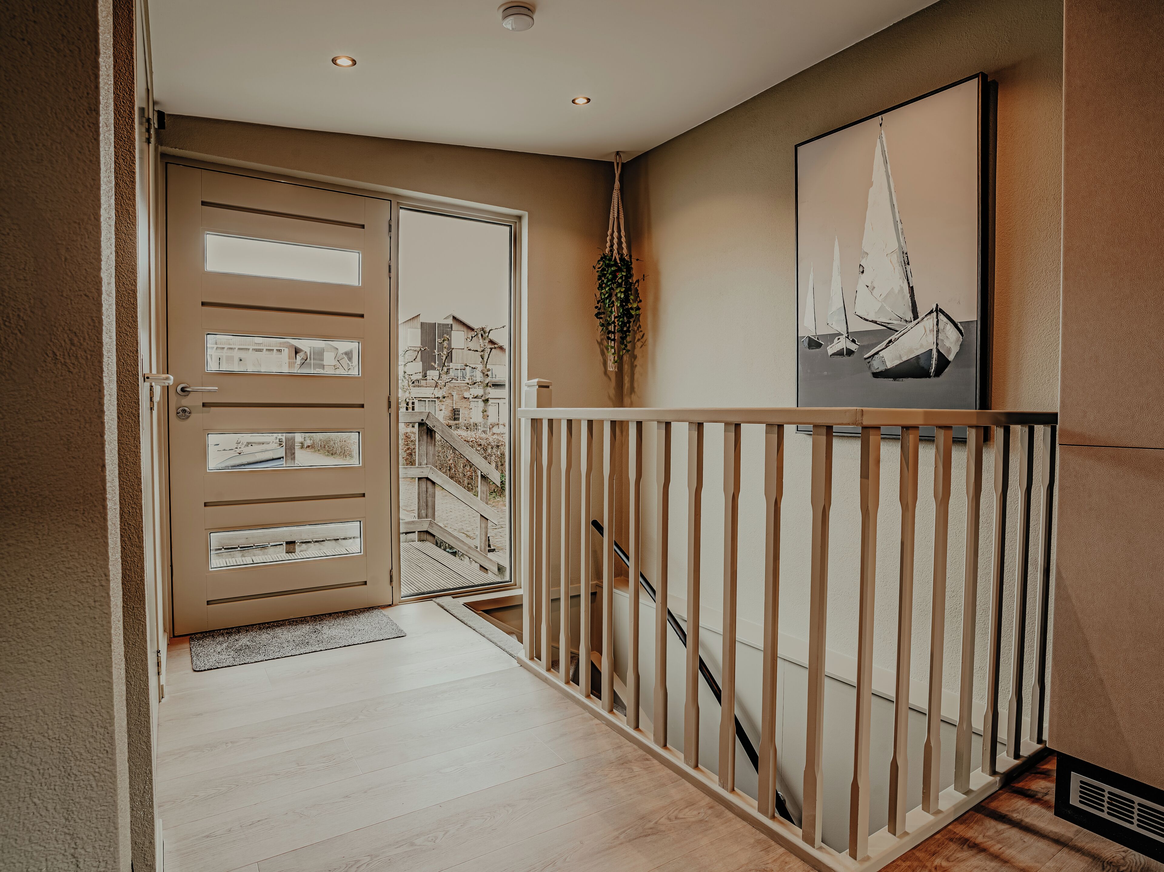 Een gang met een houten trap, een beigekleurige deur met ruitpanelen en een lijst met foto’s van zeilboten aan de muur.