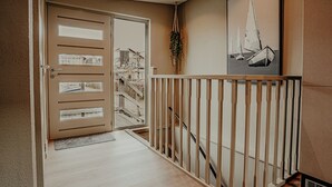 Un pasillo con escalera de madera, una puerta color beige con paneles de vidrio y un cuadro con barcos de vela en la pared.
