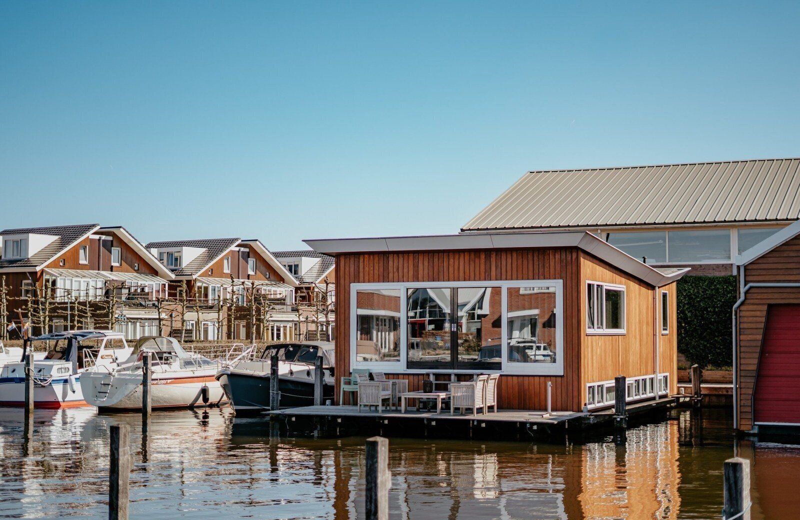 Een moderne houten woonboot afgemeerd in een jachthaven, met boten, een heldere lucht en een rustig wateroppervlak.