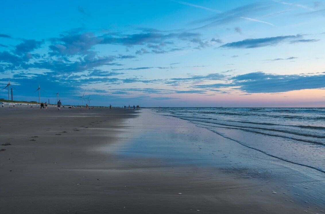 Een strand met een lange zandvlakte, enkele mensen die wandelen en windturbines in de verte.