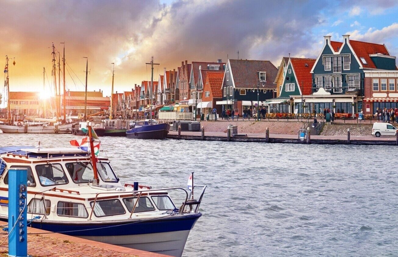 Een watergezicht met aangemeerde boten, een rij traditionele gebouwen en een ondergaande zon op de achtergrond.
