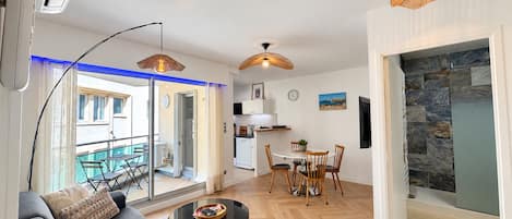 Una sala de estar moderna con un sofá, una mesa de centro y un comedor con sillas. Se observa una cocina al fondo, un balcón con mesa y sillas, y una pared de piedra.