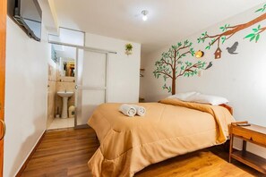 Un dormitorio con cama, suelo de madera, televisor de pared y baño con lavamanos. En la pared, un mural con motivos de árboles.