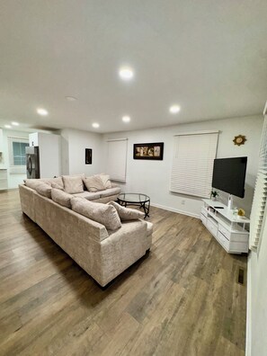 A living room with a large beige sofa, a glass coffee table, a flat-screen TV, and a white entertainment unit.