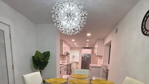 A dining area with a glass table, four chairs, a chandelier, and a kitchen in the background.