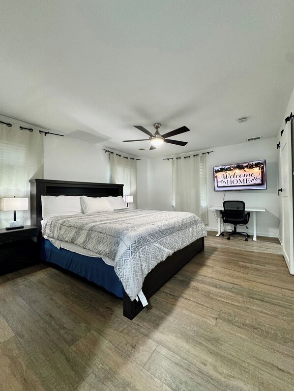 A bedroom with a bed, a desk, a chair, a ceiling fan, and a TV displaying 'Welcome HOME'.