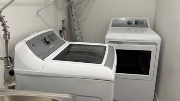 Un área de lavandería con lavadora y secadora de carga frontal, una repisa metálica con productos de limpieza y un lavabo de acero inoxidable.