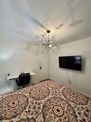 A room with a patterned carpet, a ceiling fan, a mounted TV, and a desk with a chair.