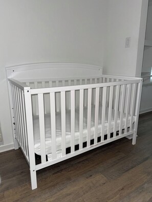 A white crib with a mattress, positioned against a wall with a wooden floor.