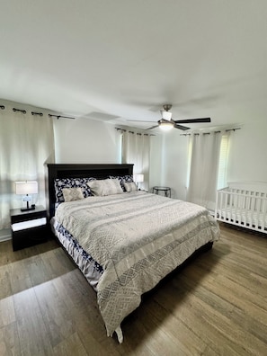 Un dormitorio con una cama, una mesita de noche, un ventilador de techo, una cuna y una ventana con cortinas.
