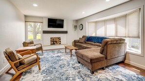 Ein Wohnzimmer mit einem Ecksofa, zwei Holzstühlen, einem Couchtisch, einem Kamin und einem großen Fenster mit Jalousien.