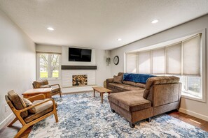 Ein Wohnzimmer mit einem Ecksofa, zwei Holzstühlen, einem Couchtisch, einem Kamin und einem großen Fenster mit Jalousien.