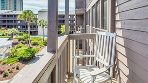 Un balcon avec une chaise blanche, une vue sur un espace aménagé avec des plantes et une piscine, et un immeuble à plusieurs étages en arrière-plan.