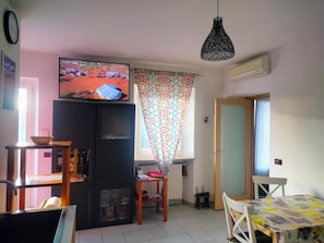 Ein Wohnzimmer mit Fernseher, einem Esstisch, einem schwarzen Schrank, einem gemusterten Vorhang und einer Pendelleuchte.