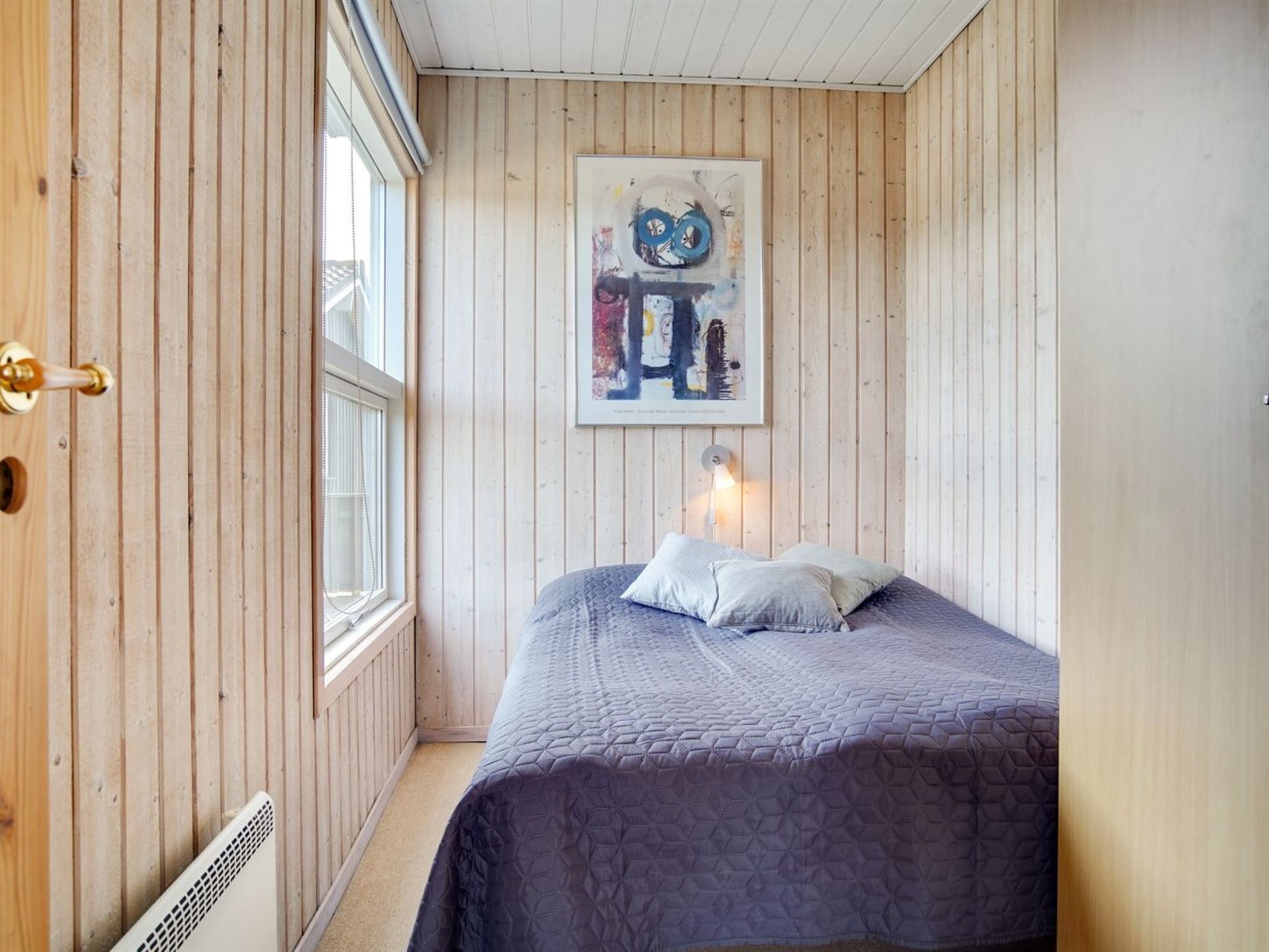 Une pièce aux murs recouverts de lambris en bois, un lit avec une couverture matelassée, une fenêtre et un tableau accroché au mur.