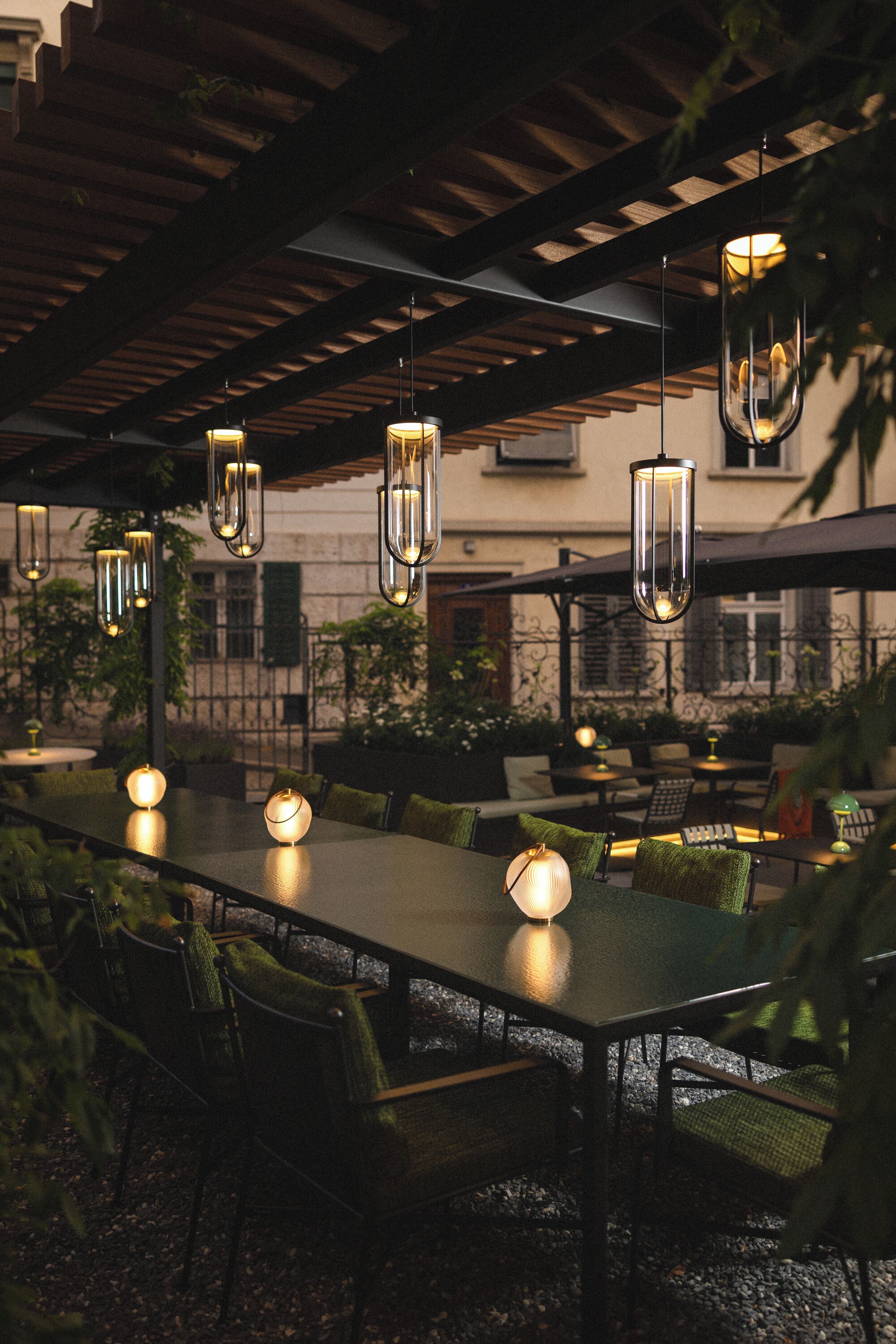 Un área de comedor al aire libre con una mesa larga, sillas, lámparas colgantes y una estructura de pérgola.
