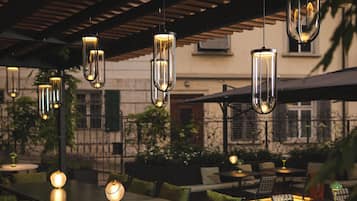 Un área de comedor al aire libre con una mesa larga, sillas, lámparas colgantes y una estructura de pérgola.