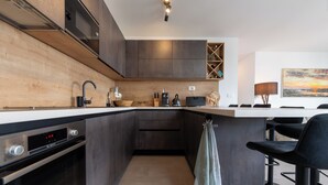 A modern kitchen with dark cabinetry, a built-in oven, a coffee maker, and a bar area with stools. - Crown Penthouse Čiovo (Okrug Gornji)