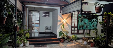 A covered outdoor area with steps leading to a balcony, surrounded by potted plants and a wall-mounted light fixture.