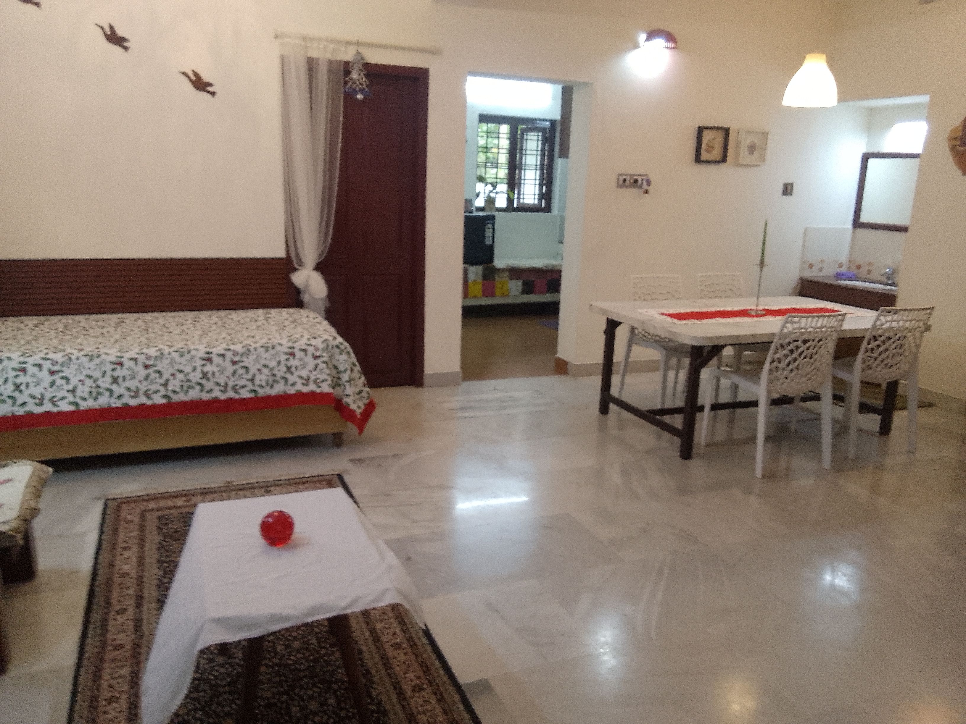 A room with a bed, a dining table with chairs, a small table with a red object, and a patterned rug.