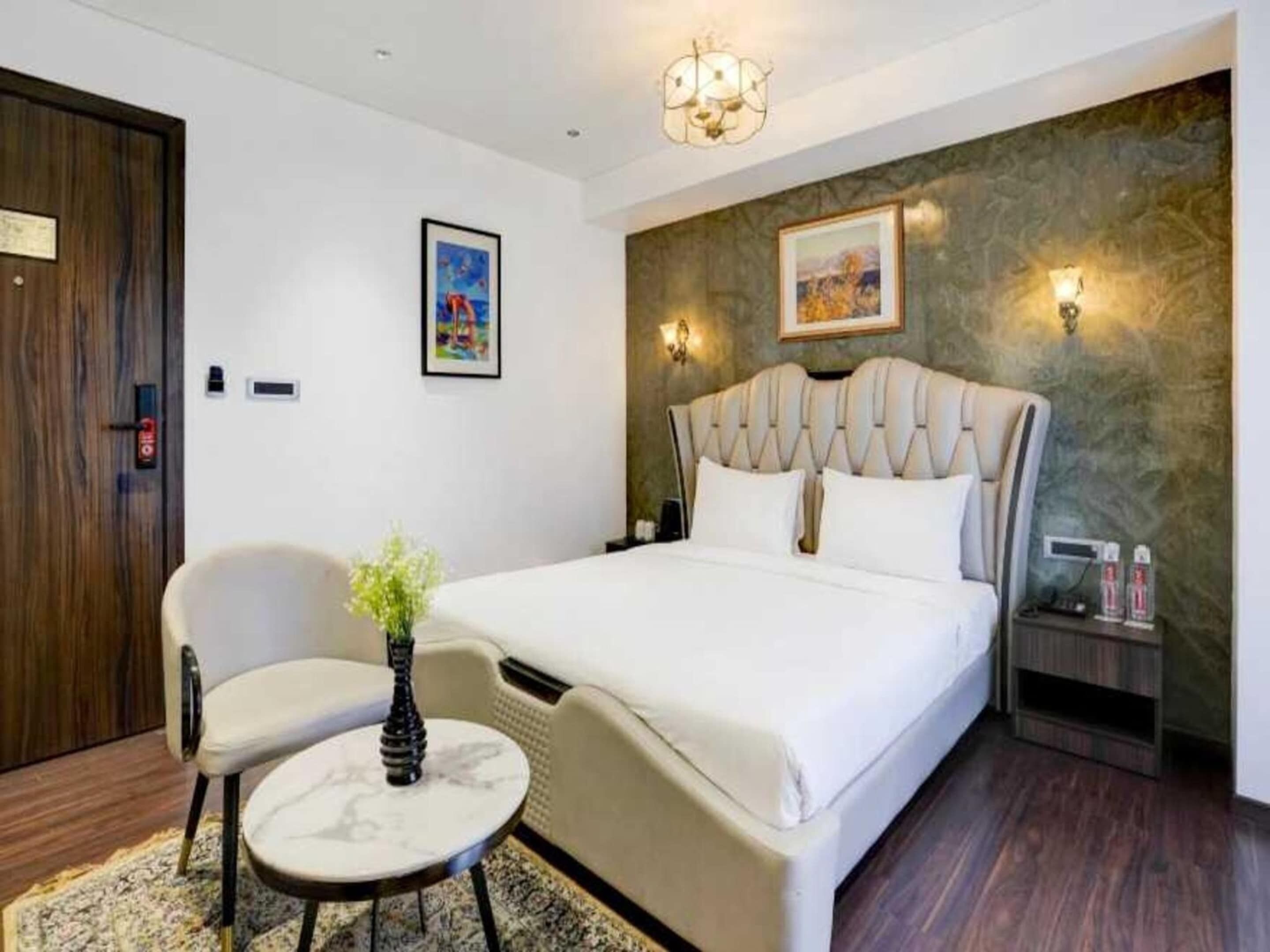 Una camera d'hotel con un letto, due poltrone, un tavolino, un vaso con fiori, un quadro appeso e un lampadario.