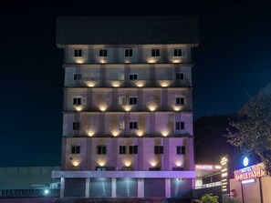 Een meerlaags hotel met verlichte ramen en balkons, een zichtbaar bord met 'Hotel Shree Yash' en een bordje 'Palette' dat een nabijgelegen gelegenheid aangeeft.
