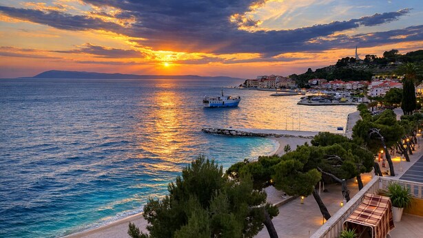 A coastal town at sunset with a boat on the water, a pier, and buildings along the shore. - Villa Sunce Podgora -studio apartment 2 (Podgora)