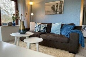 Ein Wohnzimmer mit einem braunen Sofa, zwei weißen Beistelltischen, einer Vase mit getrockneten Blumen und einem Gemälde an der Wand.