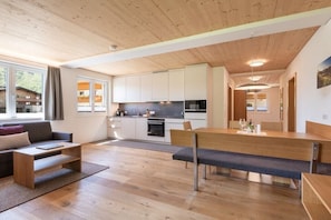 A modern kitchen with white cabinets, a dining area with a bench, and a sofa. - Rössle Apartments Type 4 (Fontanella)