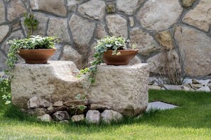Two terracotta planters with green plants on a stone pedestal in a grassy area with a stone wall in the background.
