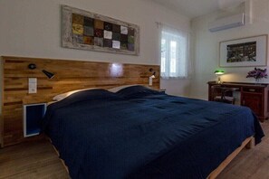 Ett sovrum med en sänggavel i trä, en säng med ett mörkblått överkast, ett nattduksbord med en lampa, en inramad tavla på väggen och ett fönster med gardiner.
