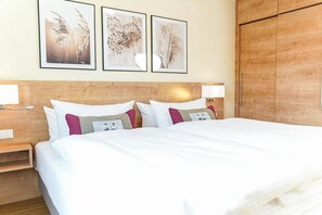Ein Hotelzimmer mit einem hölzernen Kopfteil, einem großen Bett mit weißer Bettwäsche, zwei Nachttischen mit Lampen sowie drei gerahmten floralen Wandbildern.