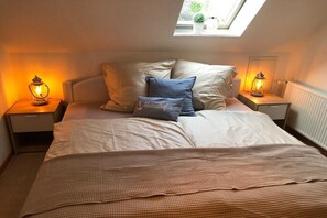 Ein Schlafzimmer mit Dachfenster, einem Bett mit karierter Decke, zwei Nachttischen mit Laternen und einem Kissen mit dem Schriftzug 'Heimathofen'.