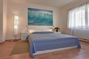 A bedroom with a bed, a nightstand, a lamp, a painting of a wave, and a window with curtains.