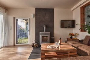 A living room with a fireplace, a sofa, a coffee table with magazines, a TV mounted on the wall, and a window with a view of the outdoors.