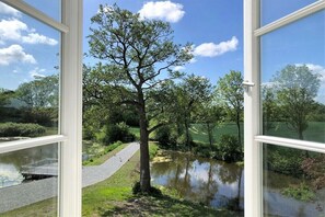Blick durch ein Fenster auf einen von Bäumen gesäumten Weg entlang eines Gewässers, im Vordergrund eine Brücke.