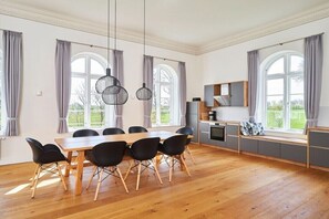 Ein geräumiger Essbereich mit Holztisch und -stühlen, eine Küche mit integriertem Sitzplatz und große Fenster mit Vorhängen.