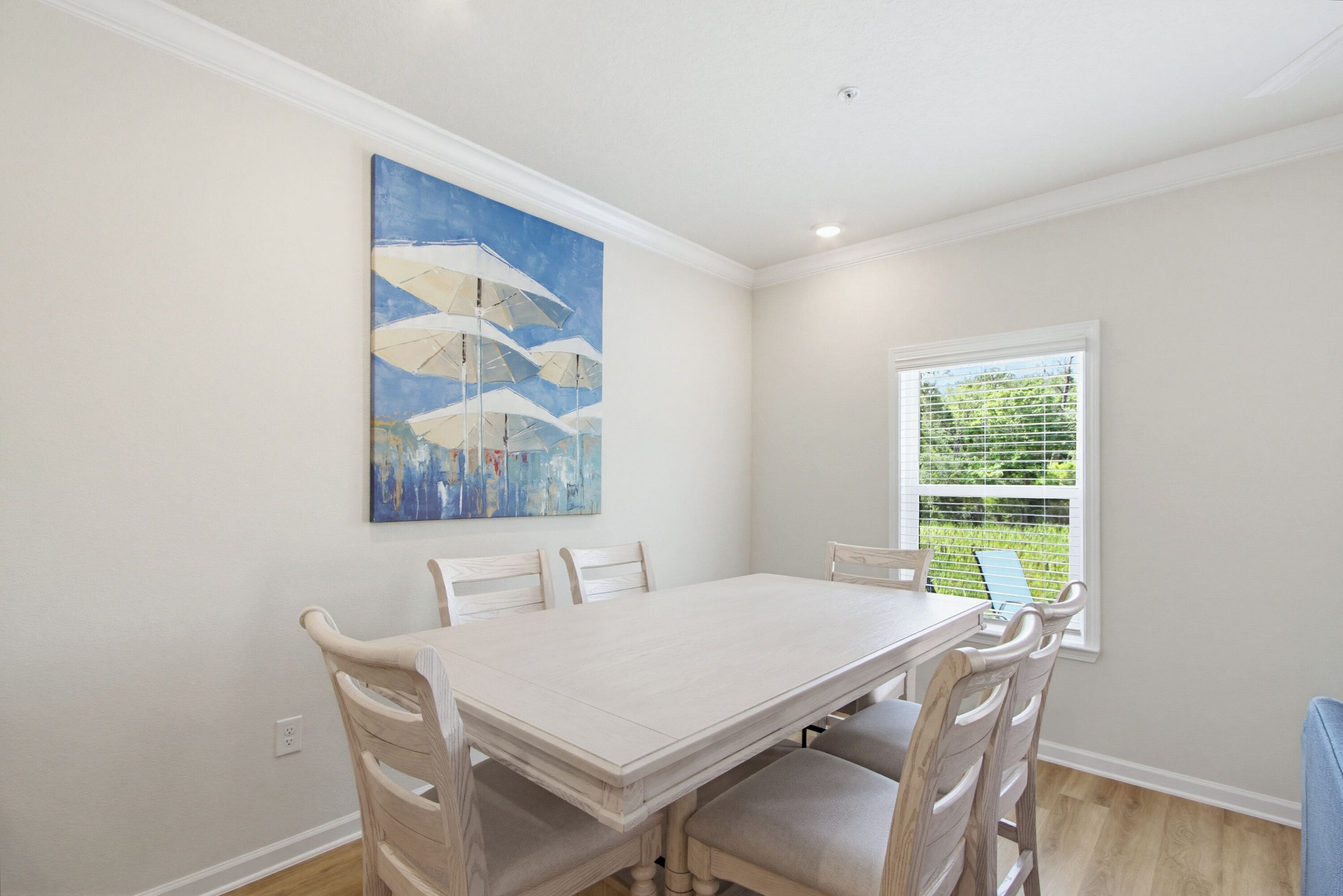 A dining room with a white table and chairs, a painting on the wall, and a window with blinds.